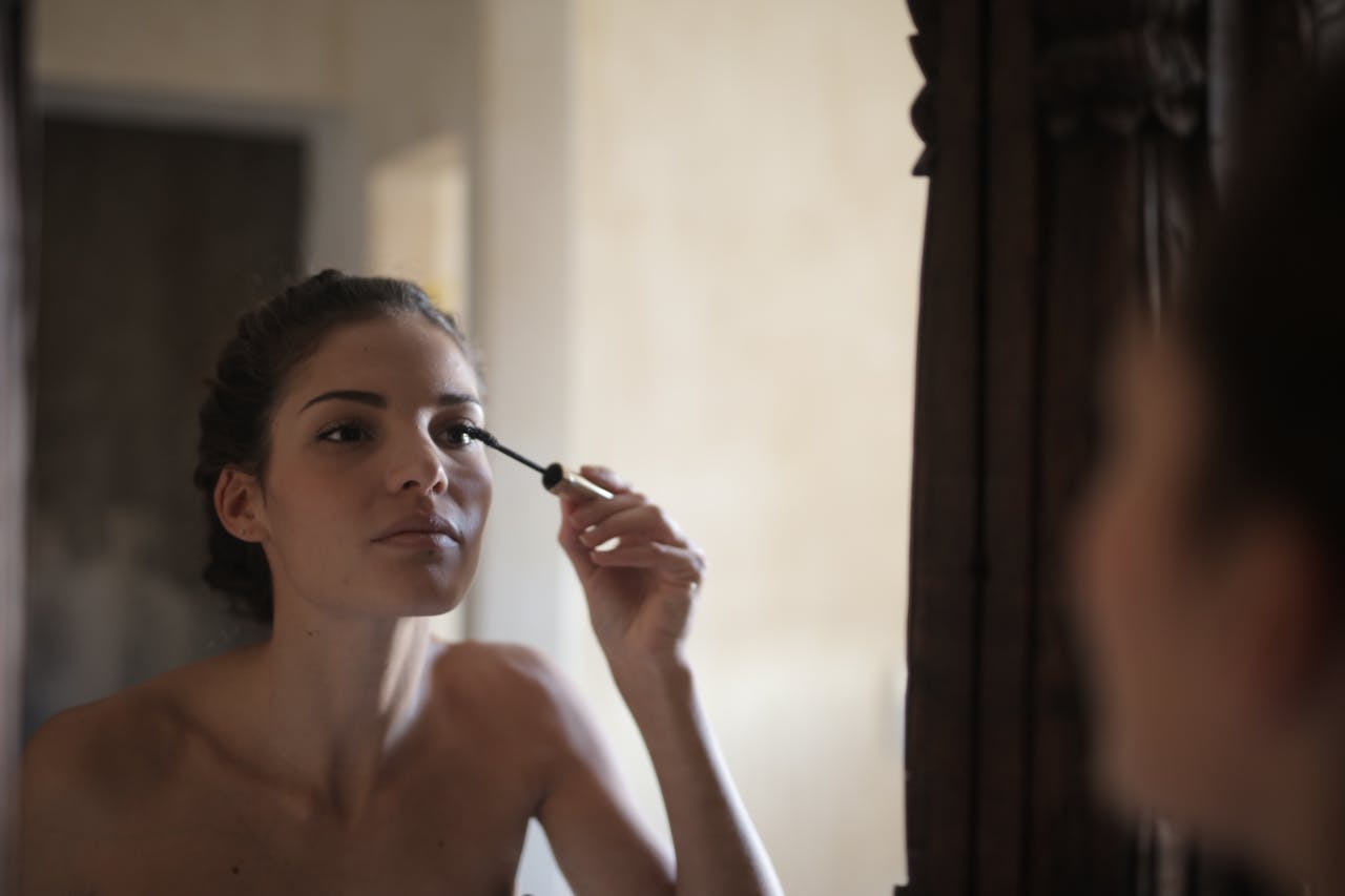 Young woman applying mascara while looking at her reflection in a mirror.