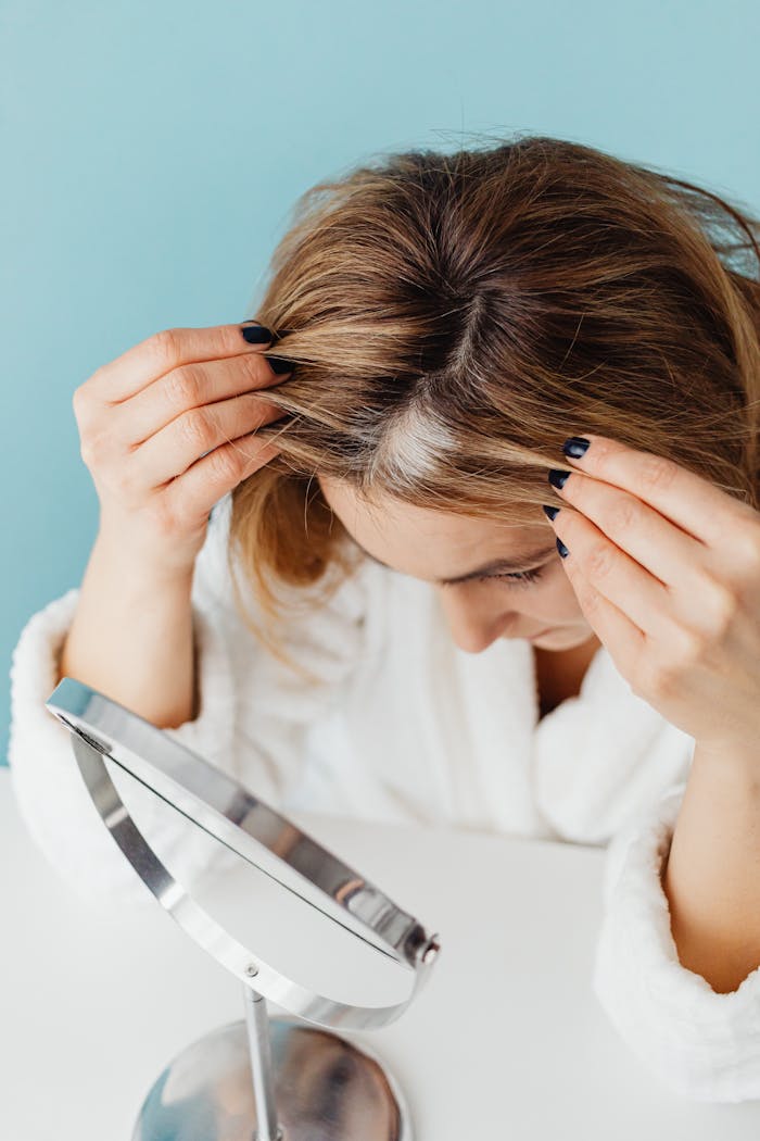 Woman in bathrobe checking grey hair roots in front of a mirror indoors.